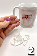 Hand holding a clear plastic heart-shaped object with a mug featuring a design of a cartoon character with a pink bow in the background.